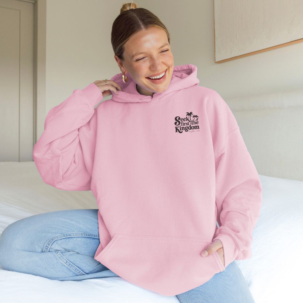 Person wearing a pink hoodie with a logo, sitting on a bed.