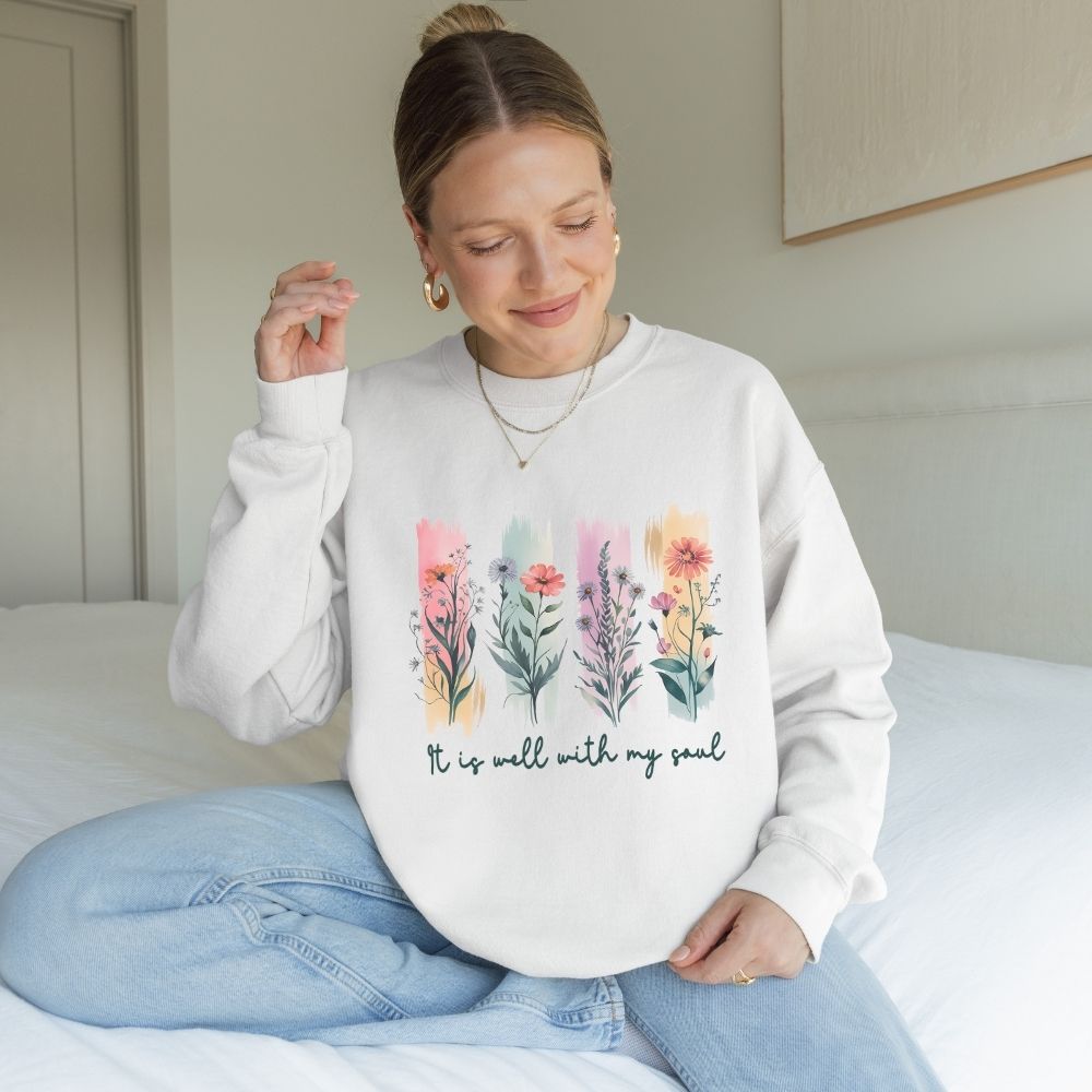Woman wearing a white sweatshirt with floral designs and text, sitting on a bed.