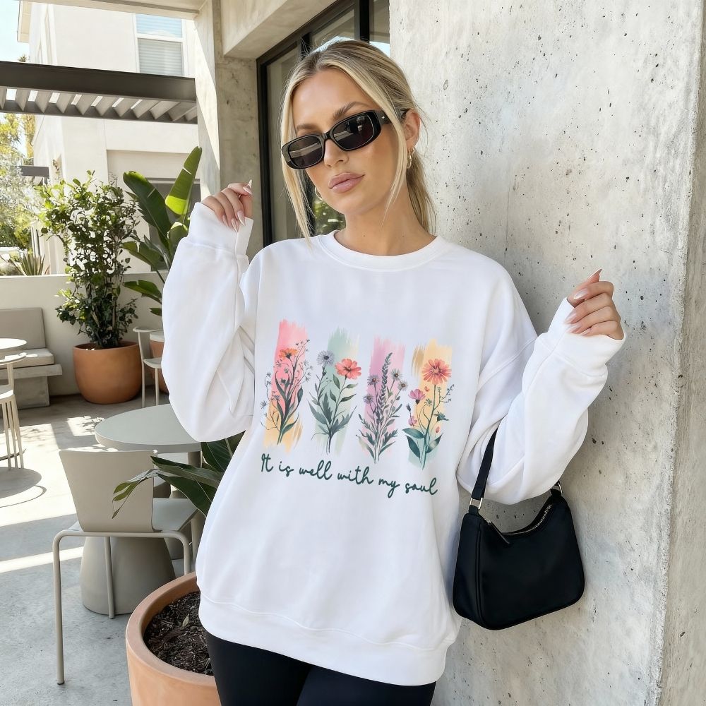 Woman wearing a white sweatshirt with floral design and text, standing against a concrete wall.