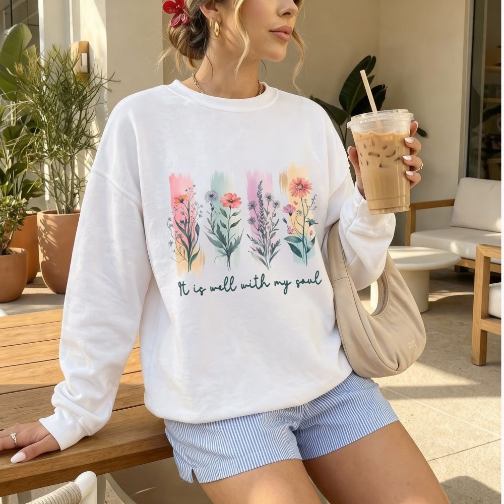 Woman wearing a white sweatshirt with floral designs and text, holding a drink outdoors.