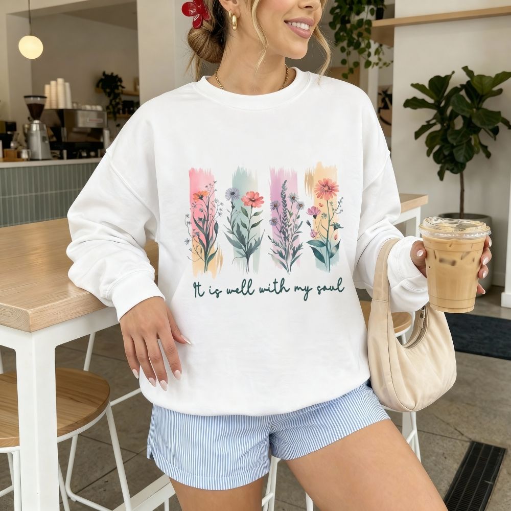 Woman wearing a white sweatshirt with floral designs and text, holding a coffee cup in a casual setting.
