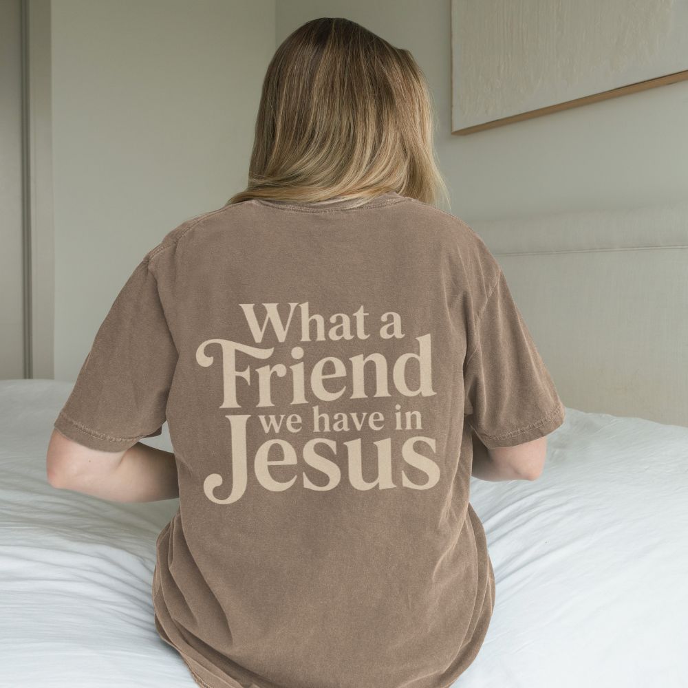 Person wearing a brown t-shirt with 'What a Friend we have in Jesus' text, sitting on a bed.