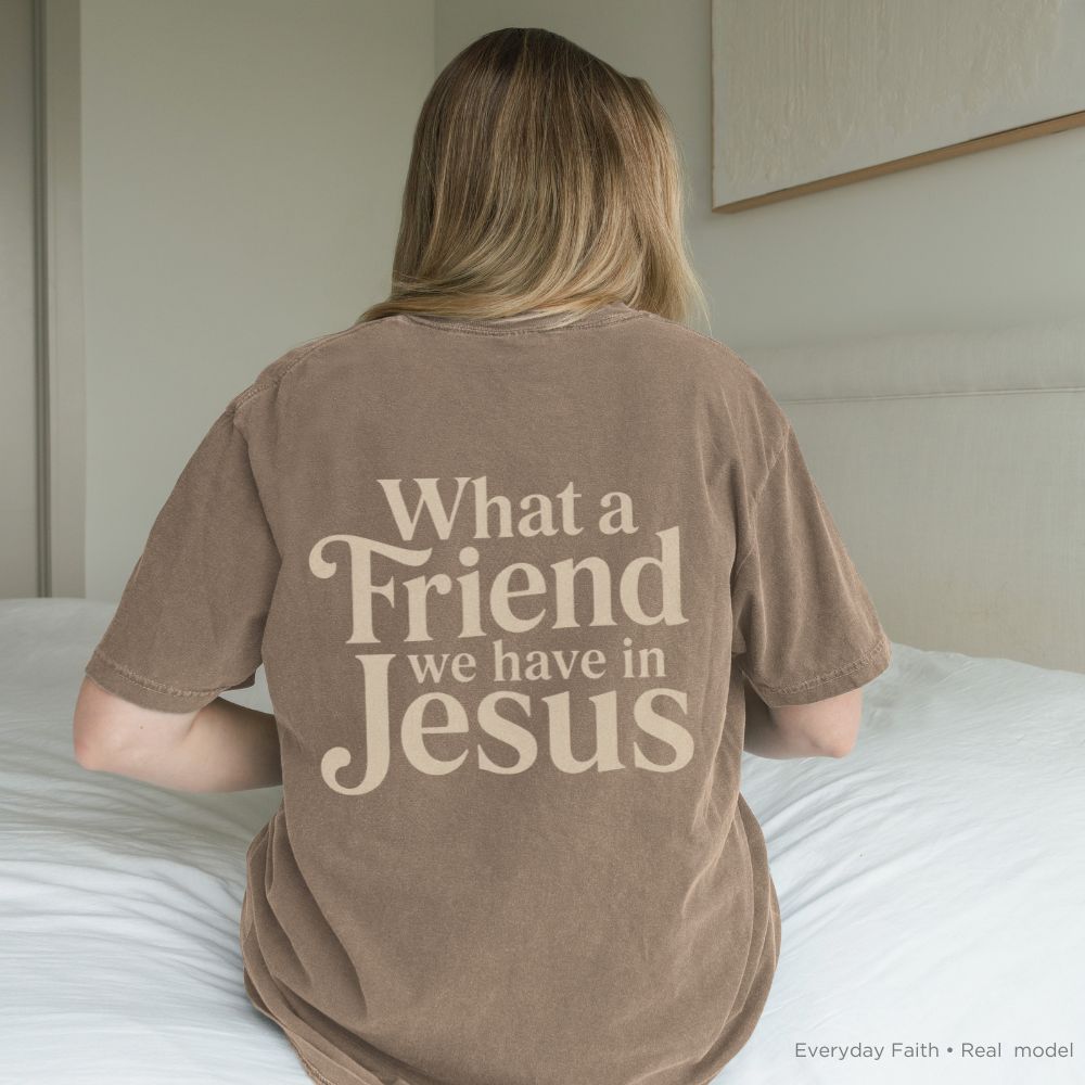 Person wearing a brown t-shirt with 'What a Friend we have in Jesus' text, sitting on a bed.