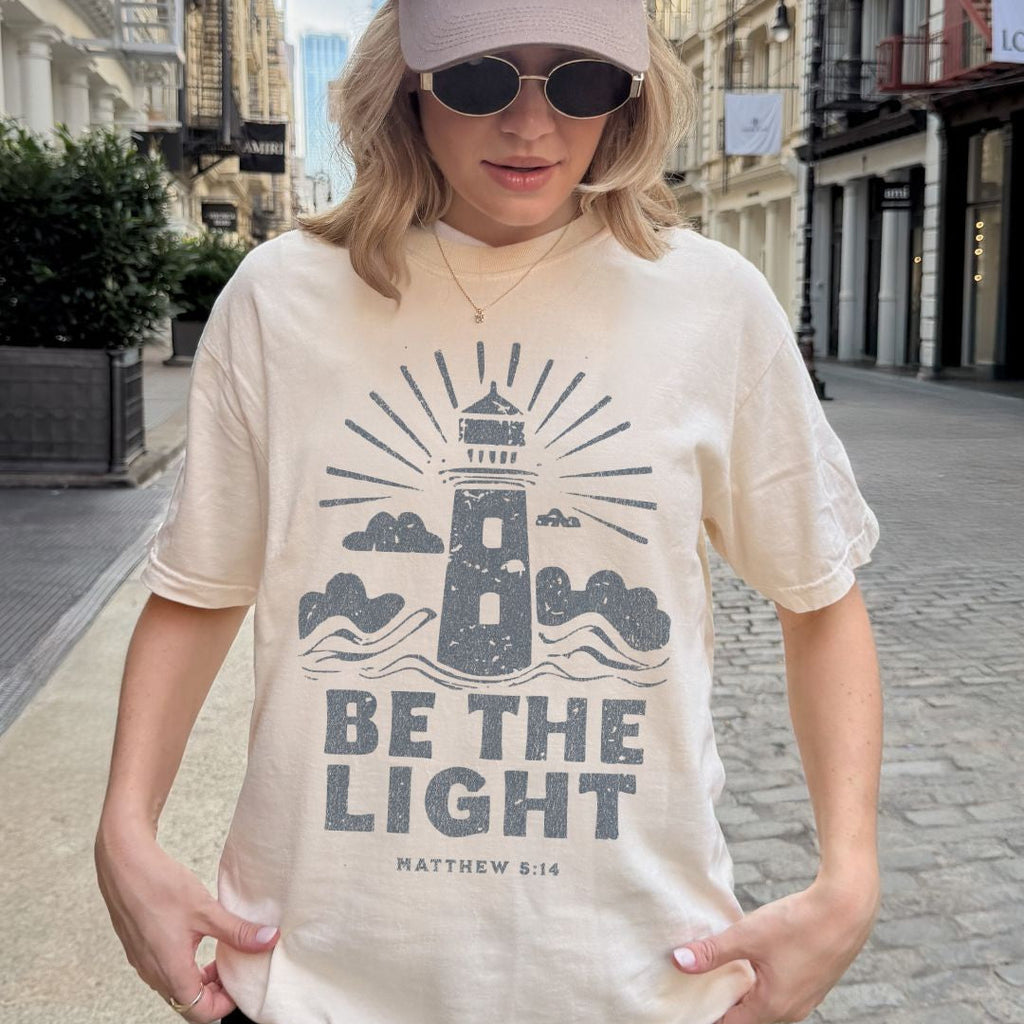Woman wearing a Be the Light Christian t-shirt featuring a lighthouse design and Matthew 5:14 scripture
