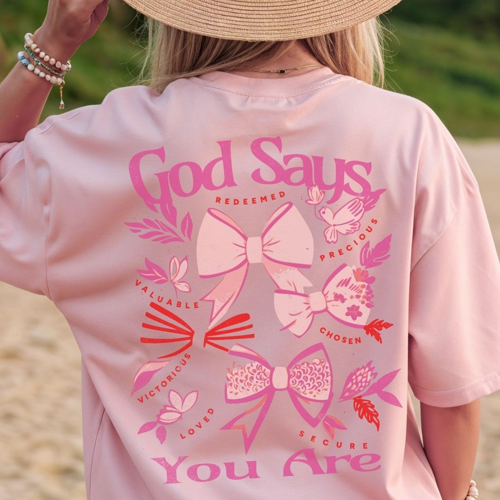 Woman wearing pink God Says You Are Christian t-shirt with bow graphics and uplifting faith affirmations on the back.