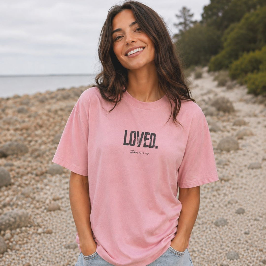 Woman wearing a pink t-shirt with 'LOVED' text on a pebbly beach.