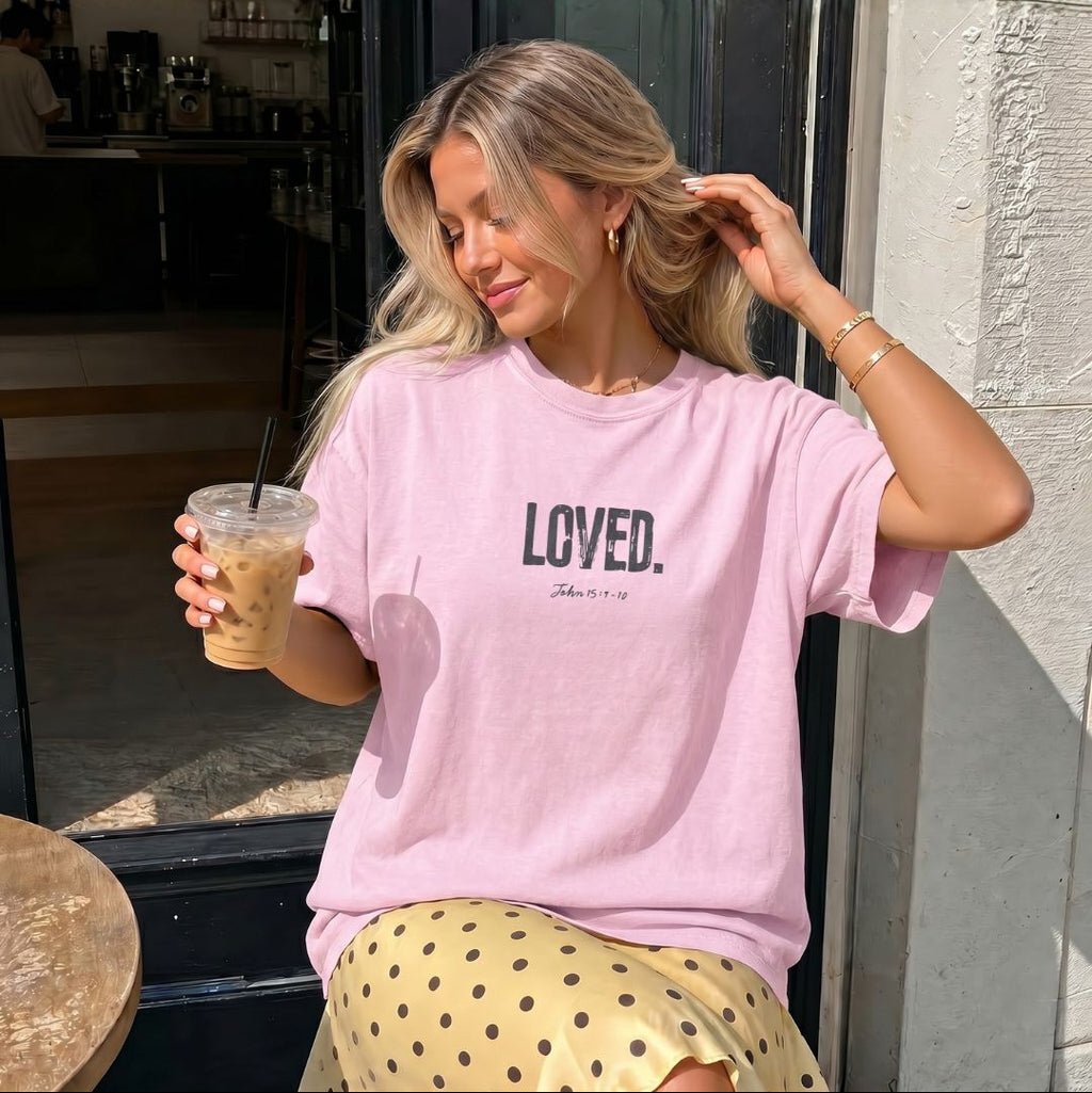 Woman wearing a pink 'LOVED' t-shirt holding a drink outside a cafe.